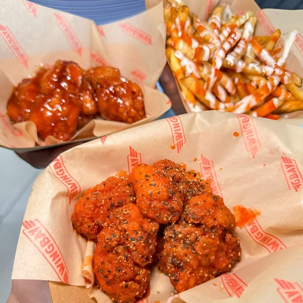 Three Wingbay baskets: two with saucy wings, one with fries drizzled with white and orange sauces.