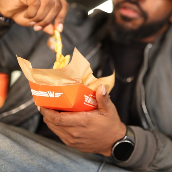 A person in a black jacket and blue jeans holds an orange Wingstop fries container, picking a fry.