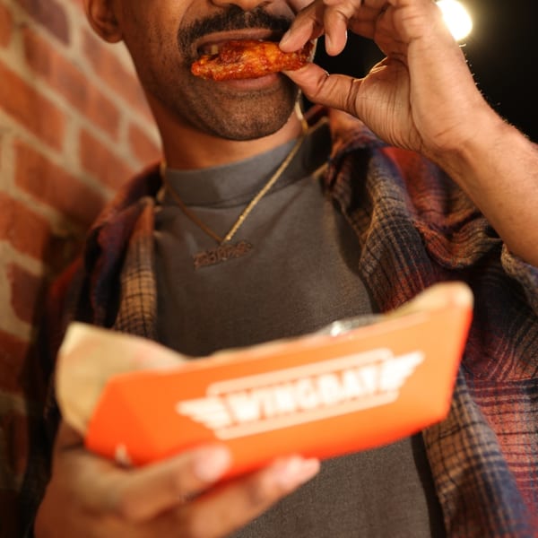 A man in a plaid shirt and gold chain eats a Wingbay chicken wing from an orange container.
