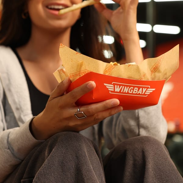 A person in a gray hoodie and jeans holds a red Wingbay fries container, eating a fry.