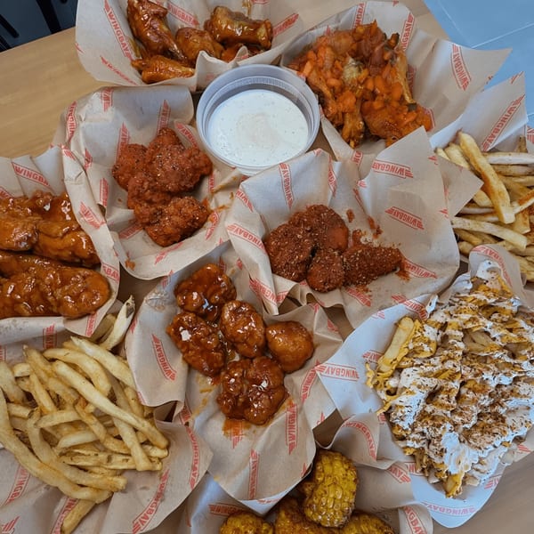An overhead view of various Wingbay chicken wings, fries, and corn in branded paper-lined baskets.