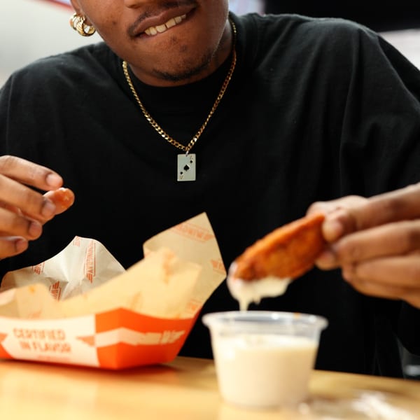 Person dipping a crispy chicken wing into a cup of white sauce, with a Wingbay box nearby.