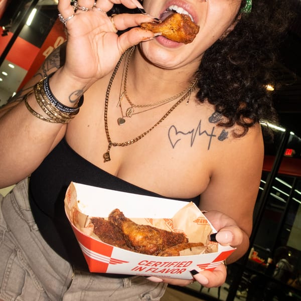 A person with curly hair and tattoos eating a chicken wing from a "Certified in Flavor" box.