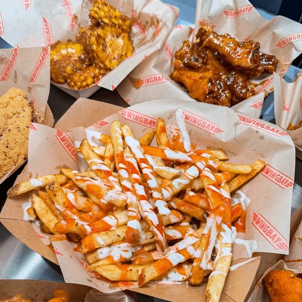Wingbay fries with white and orange sauce, corn, and wings in paper-lined baskets.